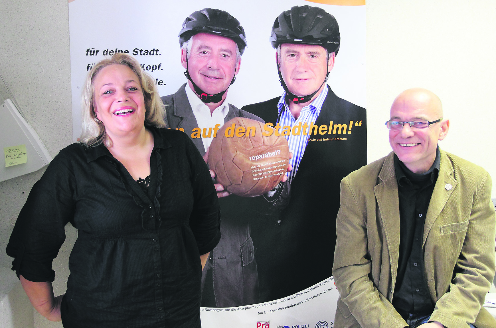Man könnte meinen, Erwin und Helmut Kremers hätten auf dem Plakat die alte Leder-Pocke gegen den Fahrradhelm getauscht. Für eine gute Sache sind die Zwillinge stets bereit, sich einzusetzen, wie hier für den Stadthelm. Das Plakat ziert natürlich auch das Büro von Christa Augustin-Sayin und Udo Böhm, denn von dem Stadthelm profitieren auch die Kinderneurologie-Hilfe und ihr Kooperationspartner, die Kinderklinik am Bergmannsheil. Foto: Gerd Kaemper