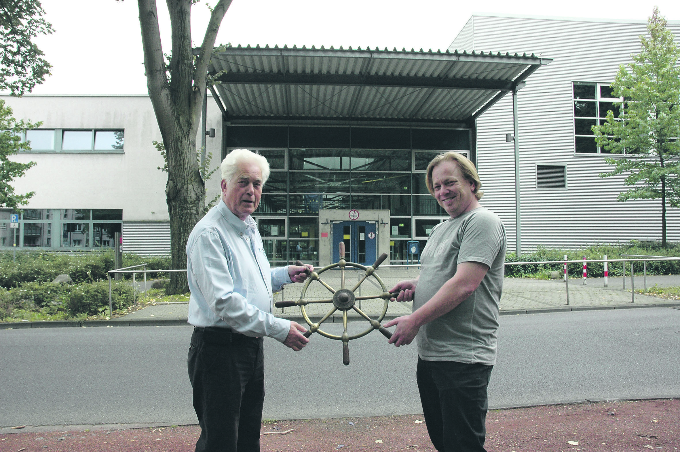 Wachwechsel: Herbert Großmann (l.) übergibt das Steuer nach 36-jähriger Amtszeit an Alex Frese, den neuen „Skipper“ des Lehrganges. Im Hintergrund das Louis-Baare-Berufskolleg, in dem der Theorie-Unterricht stattfindet. Foto: Wolf-Dedo Goldacker