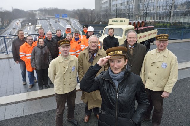 Annegret Schaber (m.), Projektleiterin Bau des Landesbetrieb Straßenbau NRW, wird neue Bierkutschermützenträgerin der Privatbrauerei Moritz Fiege. Gemeinsam mit ihrem Team, Hugo (2.v.l.) und Jürgen Fiege (4. v.l.) sowie zwei Brauereimitarbeitern posiert sie auf dem Tunnel oberhalb der Autobahn A40 in Essen. Als Projektverantwortliche hatte sie 2012 zur Sanierung der stark befahrenen Autobahn in Essen eine dreimonatige Vollsperrung anstelle einer rund zweijährigen Dauerbaustelle geplant und durchgesetzt.
