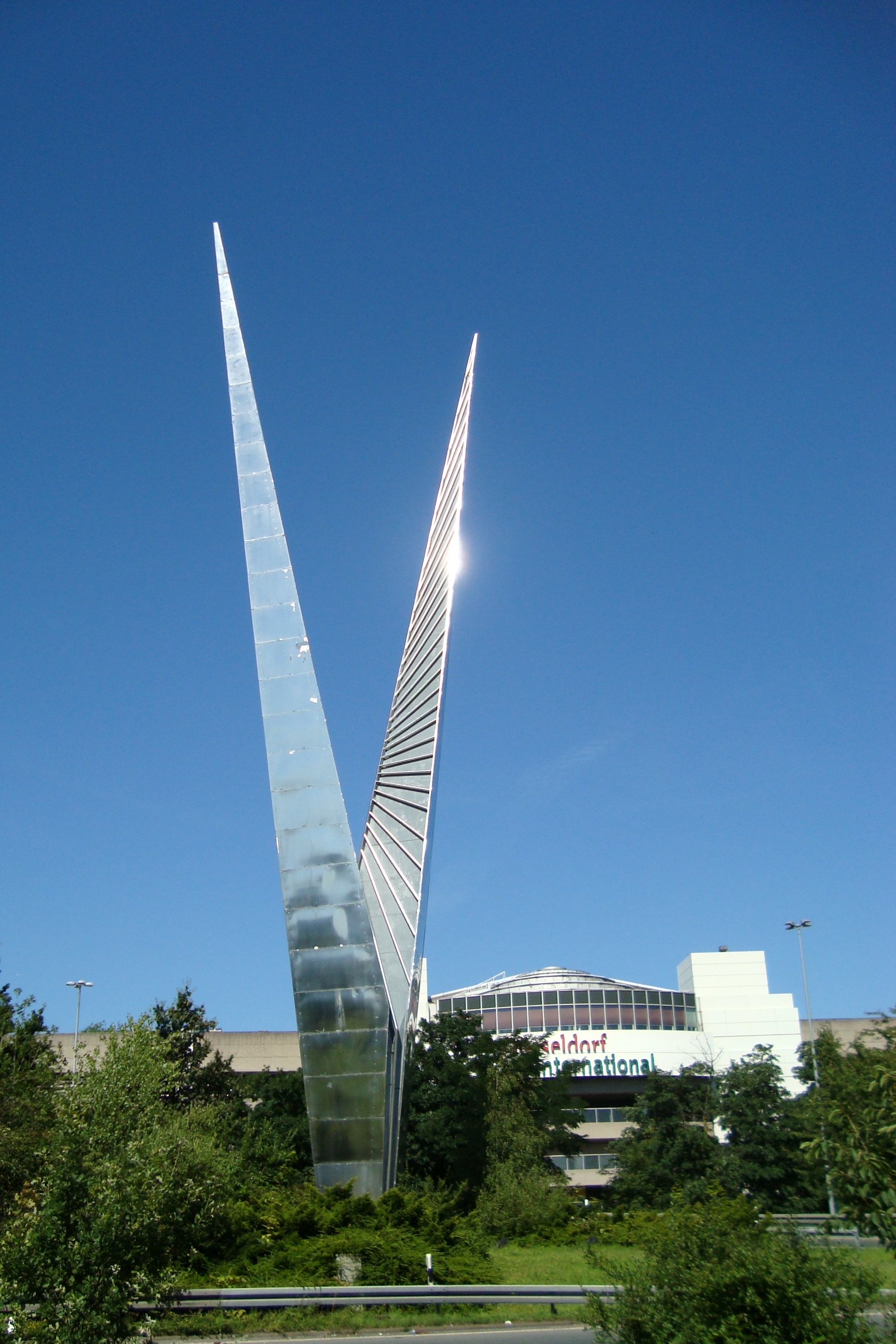 Der "Pylon" (1962) von Max Kratz, populär als "Radschläger" bezeichnet, stand ursprünglich vor der Stadthalle auf der Fischerstraße. Jetzt begrüßt die 36 m hohe Edelstahlskulptur als Wahrzeichen des Düsseldorfer Flughafens die Besucher.