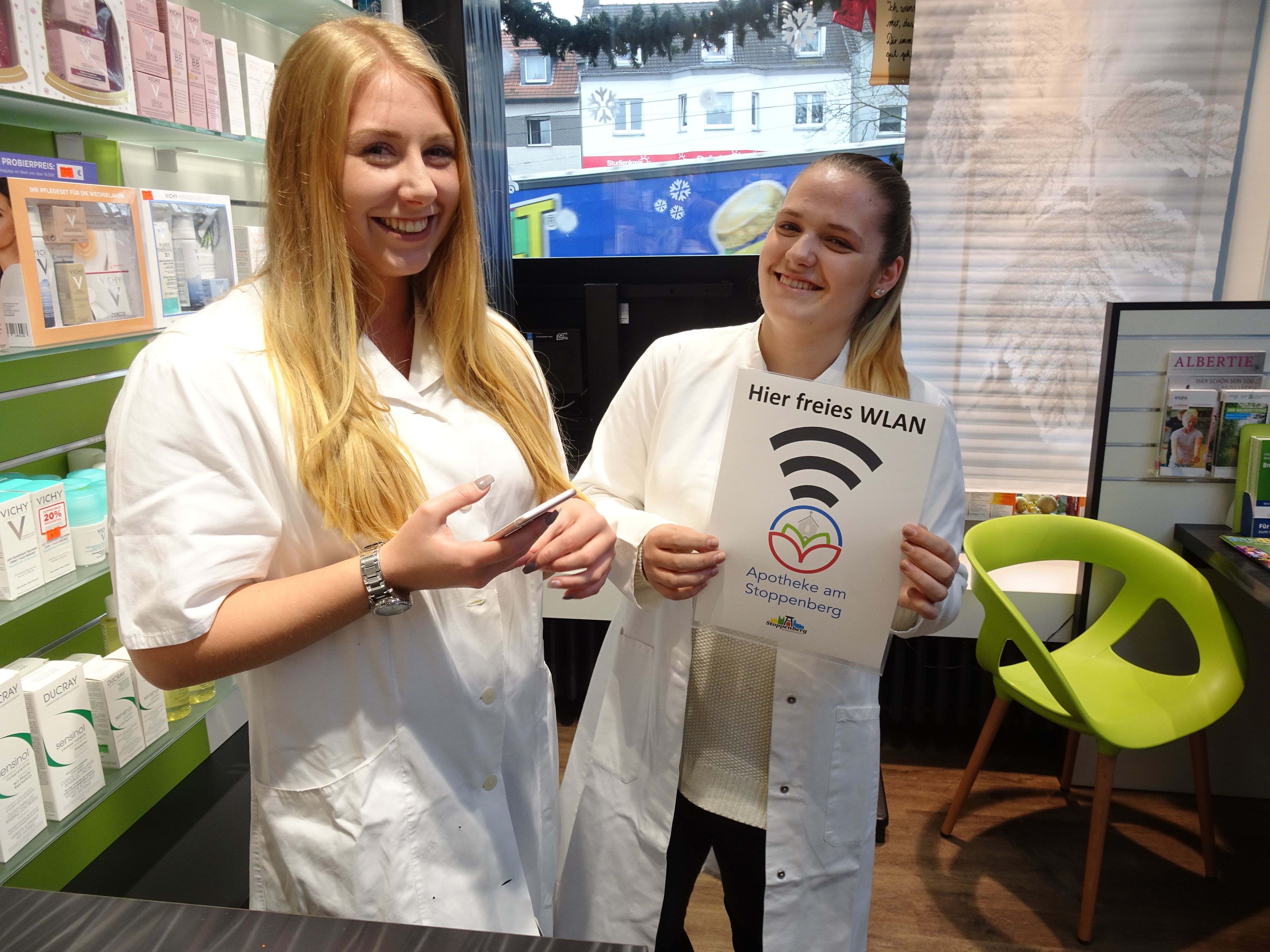 Freies WLAN gibt es jetzt in, aber auch vor der Apotheke am Stoppenberg: Charline Barkhofen (li.) und Valmire Cunaj präsentieren das Hot-Spot-Logo. Foto: Rempe