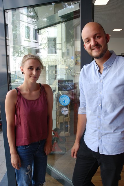 Lucy Teske und Stefan Alscher neben dem blauen Refill-Aufkleber, der auf das kostenlose Nachfüllen von Trinkwasser hinweist.
