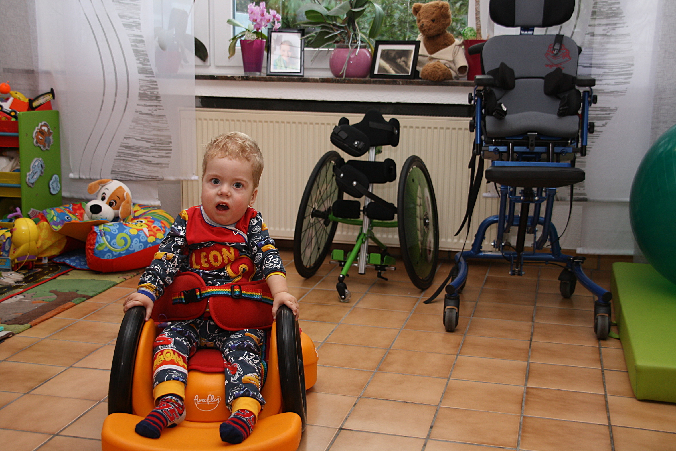 Leon fährt mit seinem Scooot. Im Hintergrund sind Stehtrainer und Therapiestuhl zu sehen.