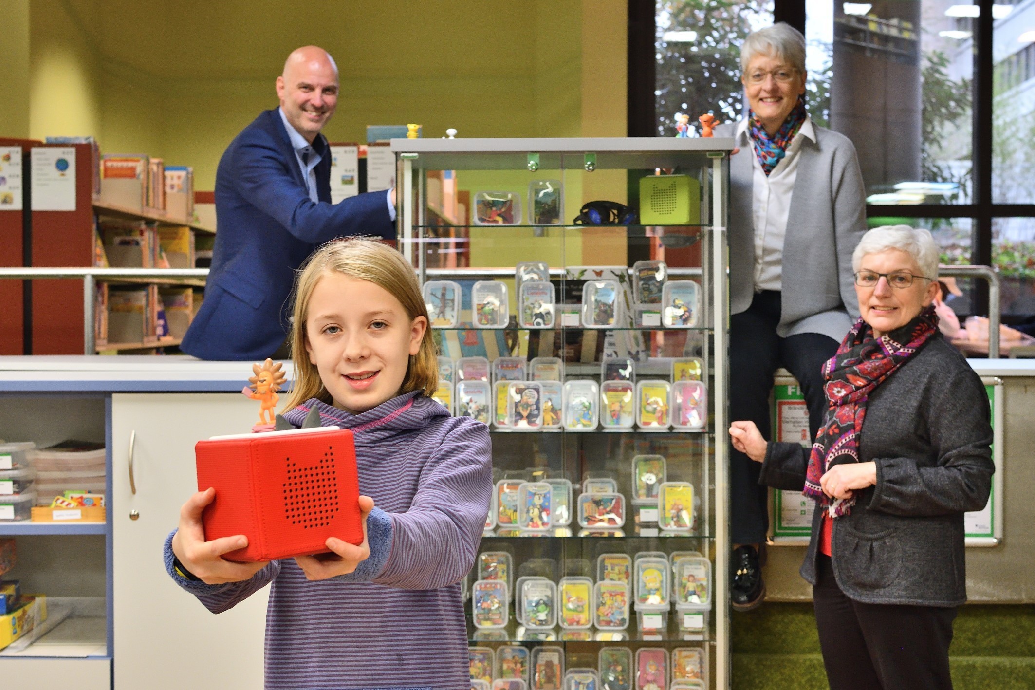 Als eine der Ersten hält Johanna eine sogenannte Toniebox in den Händen, während sich im Hintergrund Rainer Altendeitering und Ulrike Scharwächter vom Rotary-Club Bochum-Mark und Büchereileiterin Waltraud Richartz-Malmede über das neue und umfangreiche, durch den Rotary-Club finanzierte Hörspielangebot freuen.