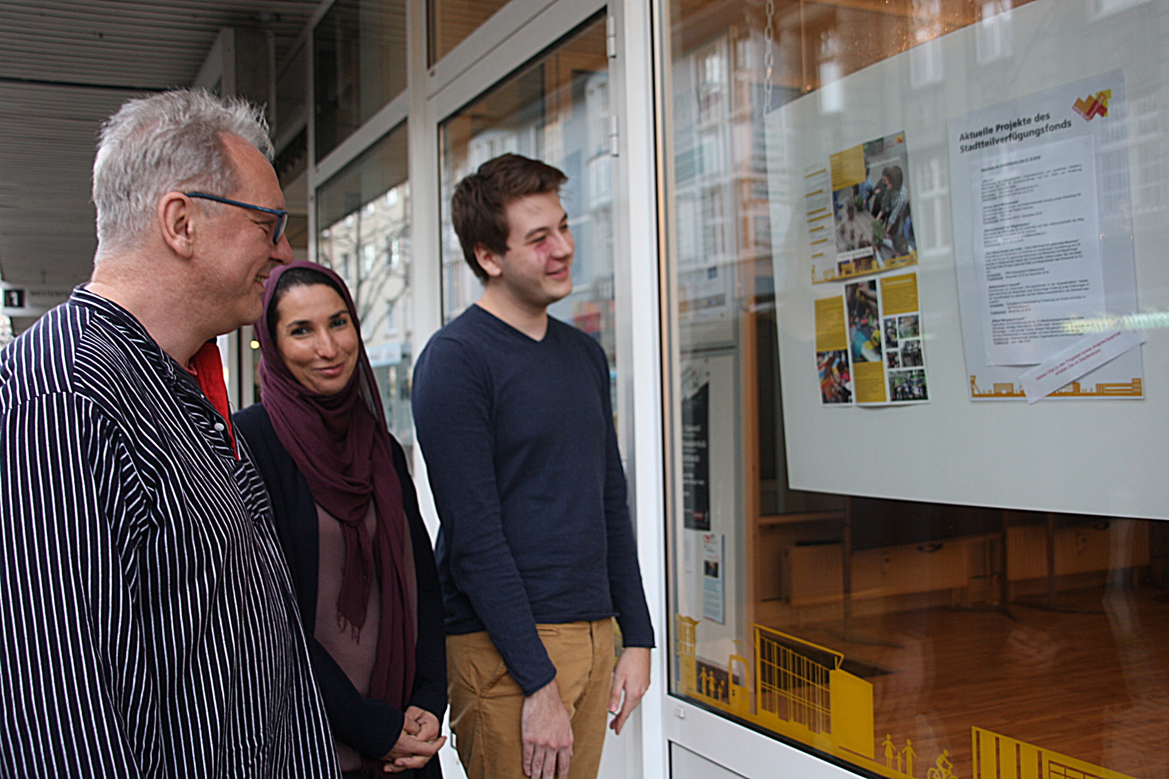 Die drei Mitglieder des Stadtteilbeirats Stephan Kreft (v.l.), Khadija Delbaz und Henri Cecatka vorm Stadtteilbüro an der Westenfelder Straße 1, in dessen Fenster die zuletzt bewilligten Projekte aushängen.