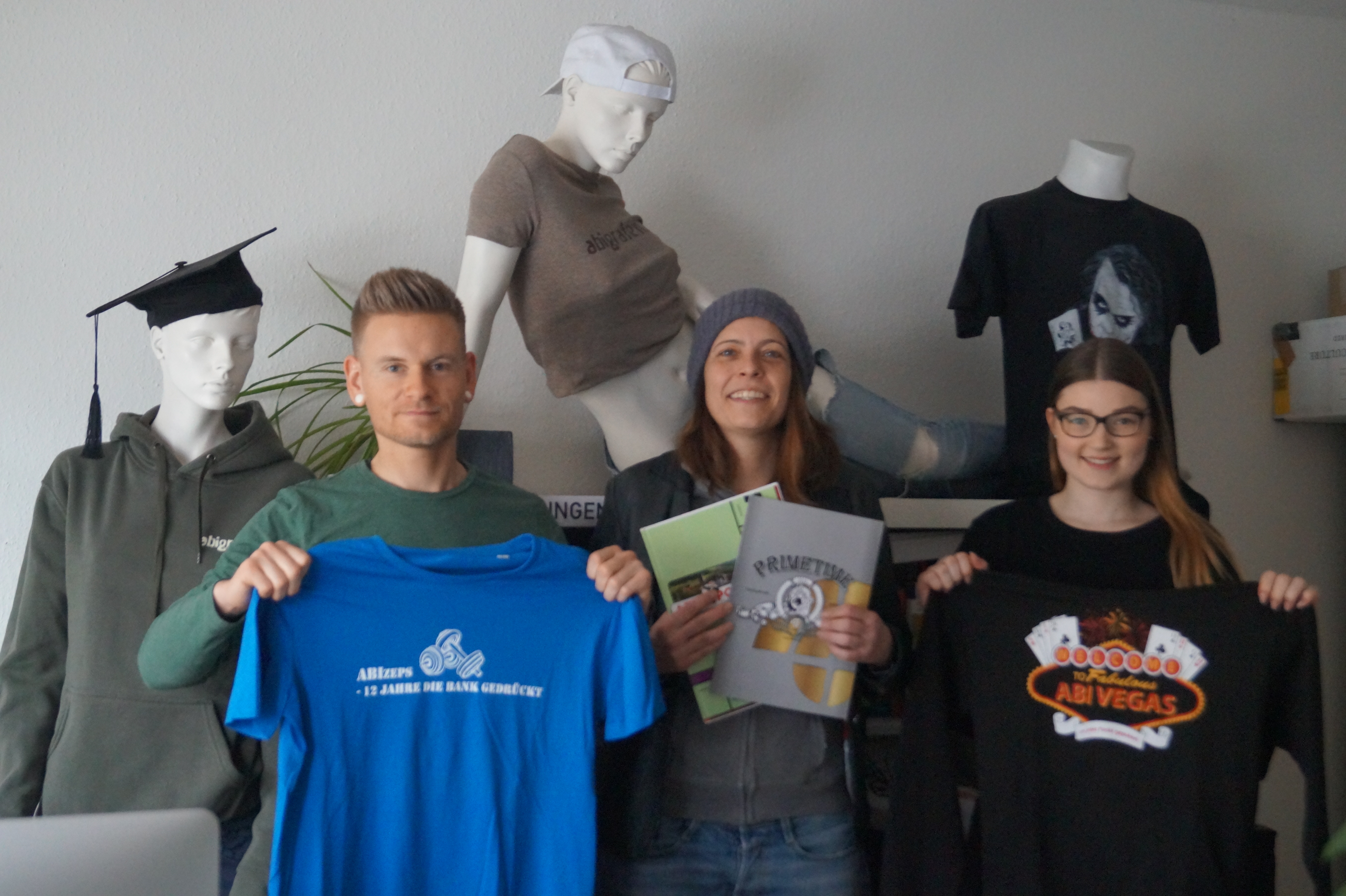 Regina Kreutner (Mitte) zeigt mit zwei ihrer Mitarbeiter, was die abigrafen.de alles so auf die Beine stellen können: Vom Shirt für den letzten Schultag über die Abi-Zeitung bis zum Doktorhut.