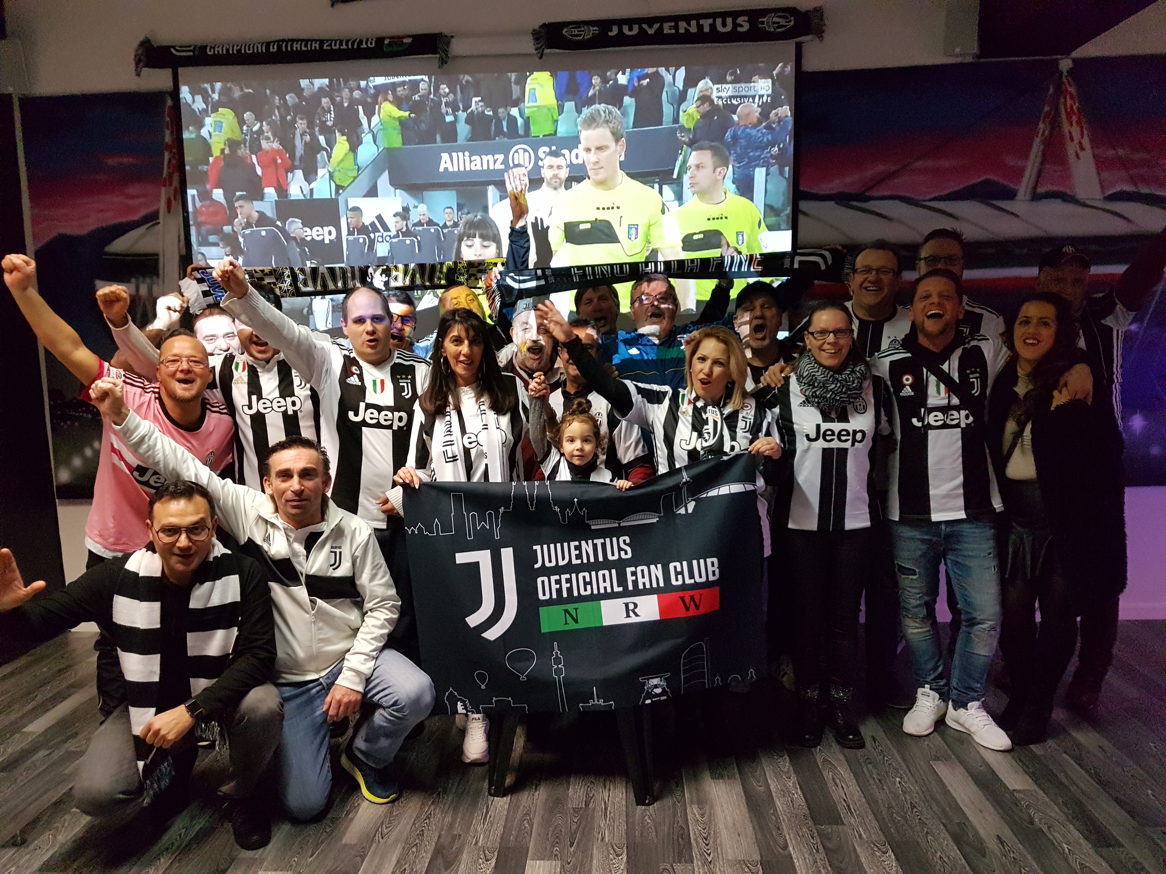 Voll bei der Sache: die Mitglieder des Juventus Official Fan Club NRW. Im Hintergrund sieht man auf der Leinwand die Mannschaften einlaufen. Eingerahmt wird diese von der riesigen Skyline des Juventus Stadium, welche sich über die gesamte Wand erstreckt.