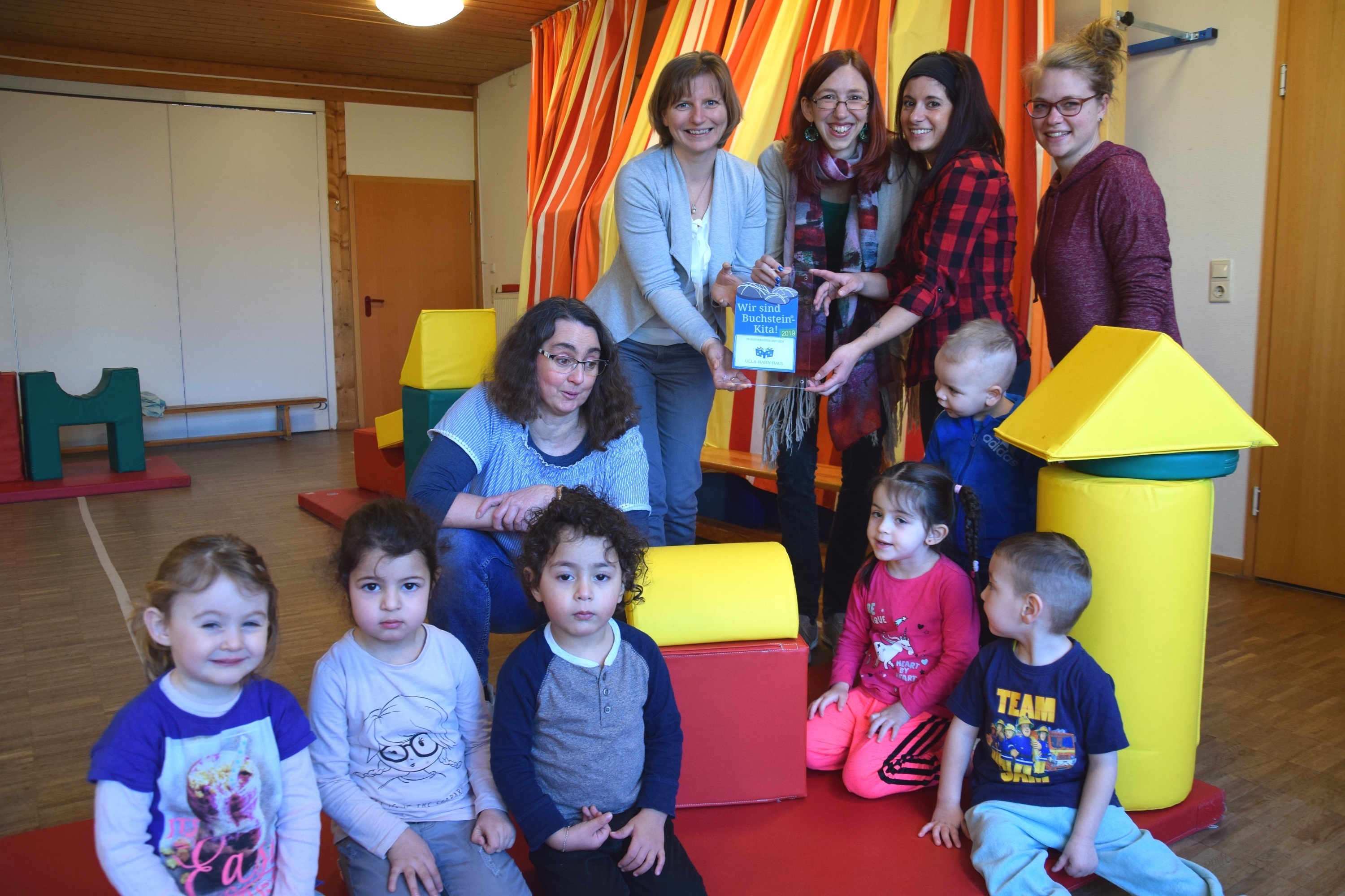 Buchstein-Kitas engagieren sich für die frühkindliche Leseförderung (von links nach rechts): Kristina Kost, Vanessa Risi-Deniz und Leiterin Kerstin Walkowiak (4.v.l.) von der evangelischen integrativen Kita und dem Mo.Ki-Familienzentrum an der Grunewaldstraße nehmen das Schild von Sonja Baumhauer (4.v.r.), Leiterin des Bereichs Bildung und Kultur und Leiterin des Ulla-Hahn-Hauses, und Projektleiterin Julia Gerhard entgegen. Foto: Birte Hauke