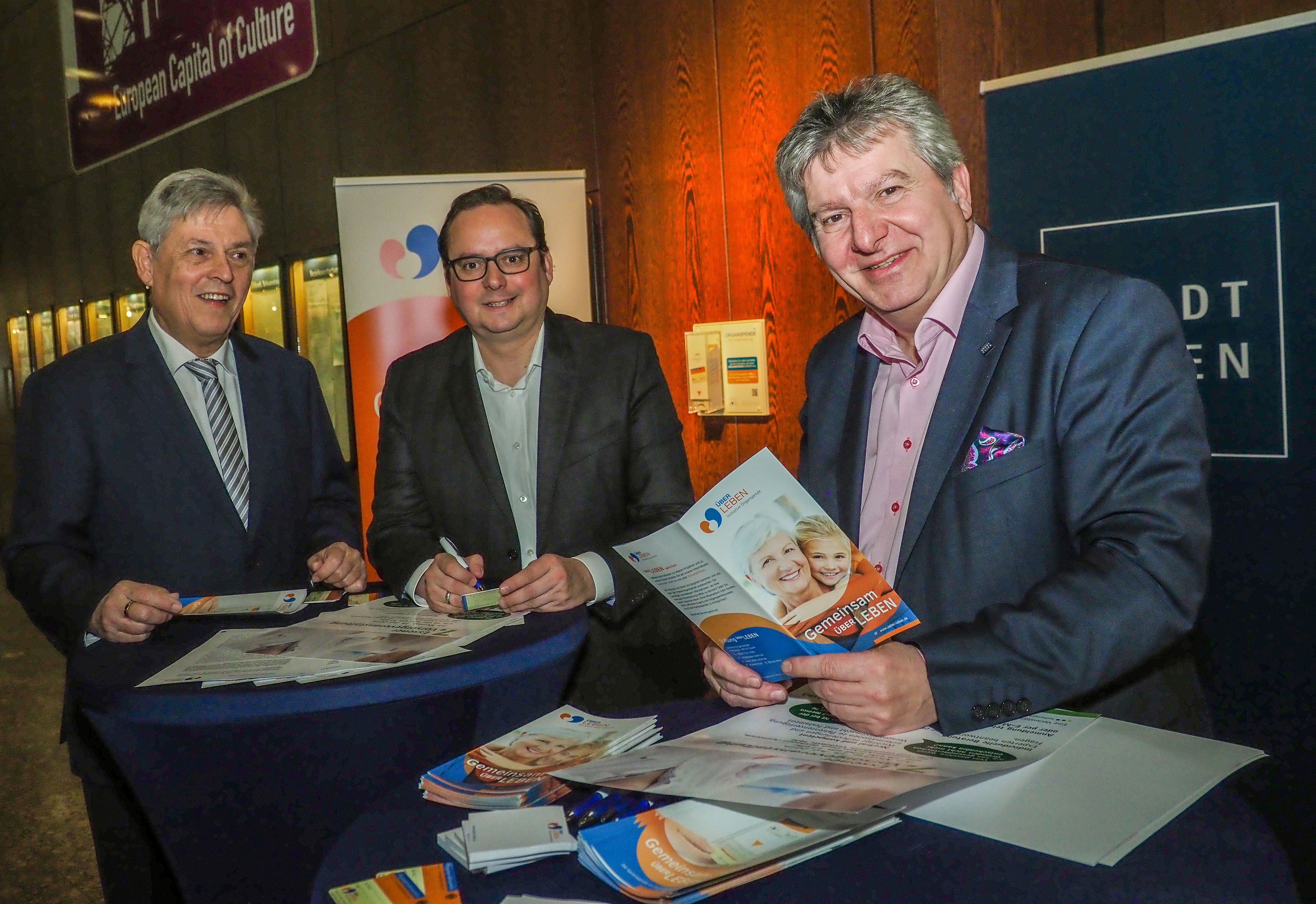Oberbürgermeister Thomas Kufen (Mitte), Prof. Dr. Karl-Heinz Jöckel, Vor-standsvorsitzender der Stiftung Über Leben – Initiative Organspende (l.) und Beigeordneter Peter Renzel am Ausweisspender im Rathaus.