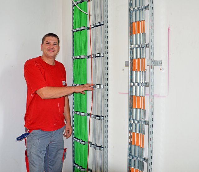 Michael Basek, Elektroniker mit der Fachrichtung Energie- und Gebäudetechnik, zeigt die verschiedenen Kabel, die für die Sicherheitsbeleuchtung (grün), den Strom (grau) und das Netzwerk (orange) verlegt werden müssen. | Foto: Maren Menke