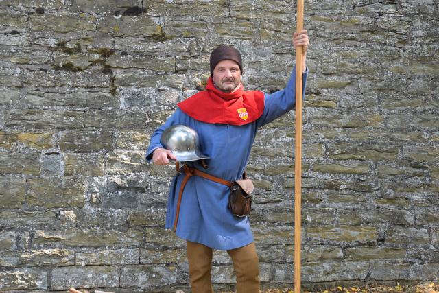 Die Pike in der Hand, den Helm unterm Arm: Auch der Lagerkoch muss für die Wache gerüstet sein.

Beim Helm handelt es sich um einen tiefgezogenen Eisenhut aus dem 15. Jahrhundert. Die Farben rot und blau waren typisch für Soldatenkleidung, da sie relativ günstig herzustellen waren. Rot wurde mit Krappwurzel gefärbt. Blau wurde mit dem Farbstoff Indigo aus dem heimischen Färberwaid gefärbt. Den Farbton der Beinlinge erreicht man mit Zwiebelschalen oder Kamille, oder einer Mischung aus beiden. Für das Braun der Mütze verwendete man die grünen Schalen der Wallnuss zum Färben.