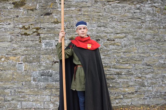 Das rote Kleidungsstück mit dem Kamener Stadtwappe ist eine sogenannte Gugel. Die ab dem Hochmittelalter nachweisbare, kapuzenartige Kopfbedeckung wurde von sowohl Männern als auch Frauen getragen und ist höchstwahrscheinlich die Namensgeberin für den Gugelhupf.

Leander Gilles trägt blaue Beinlinge und eine grüne Kotte. Die Farbe Grün ist hier eine Doppelfärbung aus Färberwaid (heimische Pfanze, ergibt blau) und anschließendem überfärben mit Reseda (Pflanze, ergibt gelb). Das erzielt dann ein kräftiges Grün, welches allerdings auch teuer war - das deutet an, das der Träger des Kleidungsstücks nicht aus armen Hause stammt.

Der Radmantel war im Spätmittelalter ein Reise- und Wetterschutzkleidungsstück und zeigt, dass dort jemand auch bei Wind und Wetter seinen Dienst tut. 

Die Pike in Leanders Hand ist mit Spitze ca. 3 Meter lang und hat einen Schaft aus Eschenholz.