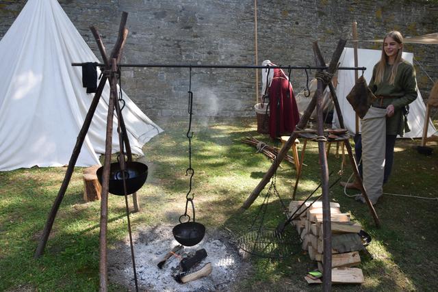 Auf der selbstgebaute Feuerstelle wird gerade das Spülwasser erhitzt. Rechts im Bild ist Leander Gilles mit seinem Köcher zu sehen. Das Zelt links im Bild ist ein Kegelzelt und ist für die im Lager dargestellte Zeit (ca. 1450) auch auf Abbildungen und Gemälden nachweisbar.