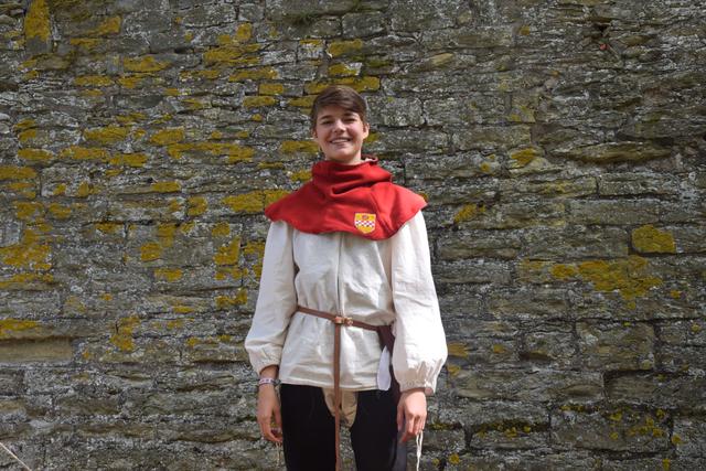 Auch hier die rote Gugel mit dem Kamener Stadtwappen. Darunter ein weißes Leibhemd und schwarze Beinlinge. Die Beinlinge wurden an der mittelalterlichen variante der Unterhose, der sogenannten Bruche, angenestelt.

Nestelschnüre waren im Mittelalter, zusammen mit Haken und Knöpfen, die übliche Verschlussart für Kleidung. (Im Bild zu sehen ist Philine Gilles)