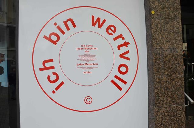 Hermann J.Kassel „stempelt“ eine Fensterfläche der Ausstellung mit seinem Slogan „Ich bin wertvoll“, den er anlässlich seiner Arbeiten rund um das Thema Menschenwürde entwickelt hat.