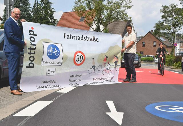 v.l. Technischer Beigeordneter Klaus Müller, Heribert Wilken, Leiter des städtischen Fachbereichs Tiefbau präsentieren Banner zur Fahrradstraße und Straßenverkehrsamtsleiterin Monika Werwer auf dem Fahrrad an der Schützenstraße. Foto: Stadt Bottrop