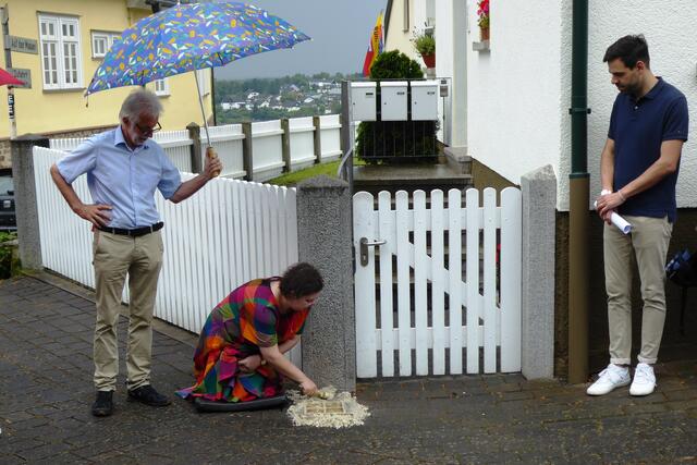 Hannah Mills legt die Stolpersteine ihrer Battenberger Vorfahren frei. Links im Bild Norbert Becker, der jetzige Eigentümer des Hauses Hauptstraße 54. Rechts beobachtet Ortsvorsteher Felix Alexander Horsel die Freilegung.