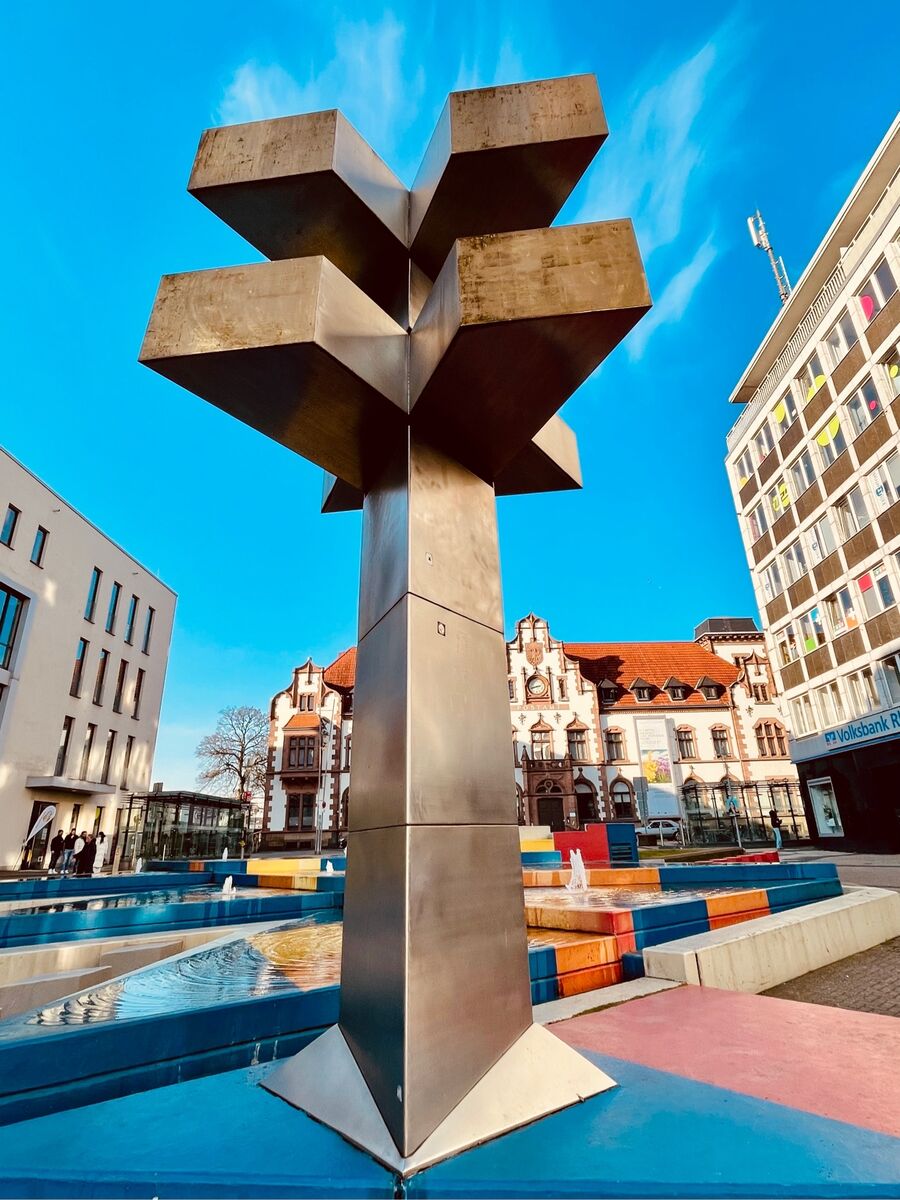 Vor dem Medienhaus erhebt sich aus einer aluminiumfarbenen Rautenform eine 6,5 Meter hohe „Säule“ . ein Körper aus Edelstahl. Diese wird aus drei liegenden Buchstaben V gebildet; das mittlere um 180 Grad zu den beiden anderen gedreht. Die V-Form leitet sich vom ursprünglichen Namen des Platzes ab: Viktoriaplatz. Entworfen hat diese Säule Otto Herbert Hayek (1927-2005)  – gefertigt wurde sie bei Thyssen-Steel. Kosten der Brunnenanlage seinerzeit 450.000 € - letzte Auffrischung 17.000 €. | Foto: Mülheimer Kunstverein und Kunstförderverein KKRR