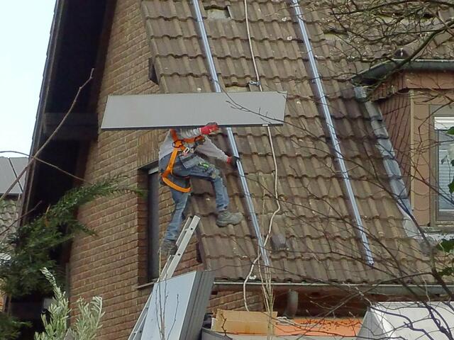 Montage einer Photovoltaikanlage - Handwerker besteigt mit Solarmodul auf der Schulter über eine unsicher angelegten Anlegeleiter das Dach eines Wohnhauses. 
Aufstiegsgerüst, Arbeits- und Schutzgerüst, sichere Arbeitsausführung = Fehlanzeige! 

