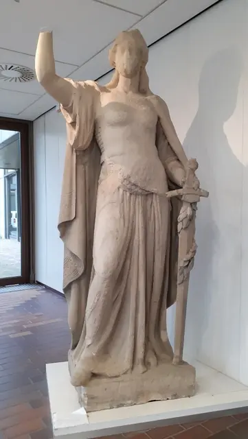 Abb. 4. „Germania“ im Foyer des Stadtmuseums, eigenes Foto (2023). 1918 Abschlag der Kaiserkrone durch Bilderstürmer, 1945 Granatsplitterschäden an Hüfte und Gürtel, bis 1981 Erosion des Gesichts und des Brustpanzers durch sauren Regen. | Foto: Eigenes Foto.