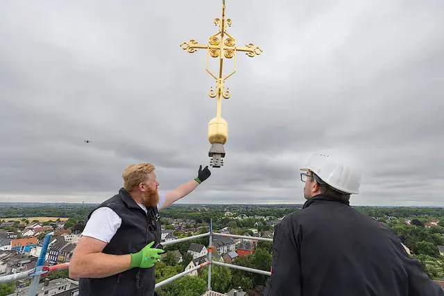 Dachdecker Sebastian Schulze (links) und Kunstschmied Rafael Jürgens nehmen das am Kranhaken schwebende Turmkreuz auf der Kirche St. Maria Magdalena in Empfang.  | Foto:  Achim Pohl | Bistum Essen